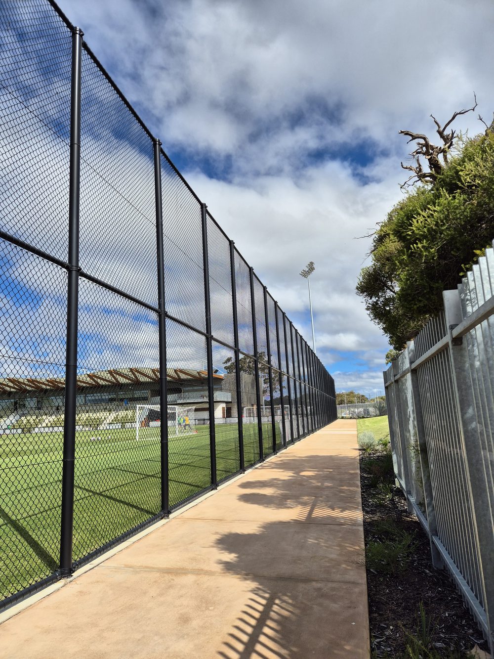 Tall chainmesh oval fencing alongside a walkway at the State Football Centre, with the grandstand behind